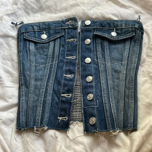 Jeanskorsett med knappar och rå kant - Säljer en snygg korsettopp i blå denim med silvriga knappar framtill, dekorativa fickor och rå, fransig nederkant. Toppen har markerade sömmar och elastisk smock baktill för skön passform. Perfekt för dig som vill sticka ut med en edgy jeanslook.