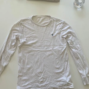 Vit långärmad tröja från Polo Ralph Lauren - Vit långärmad tröja från Polo Ralph Lauren med klassisk rund hals och den ikoniska blå broderade loggan på bröstet. Tillverkad i mjuk bomull som är skön mot huden. Perfekt för chill dagar och enkel att matcha med jeans eller shorts.