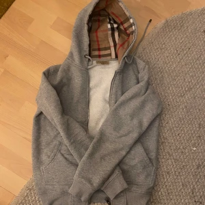 Grå hoodie från Burberry - Säljer en stilren grå hoodie från Burberry med klassiskt rutigt mönster i luvan. Tröjan har dragkedja och broderad logga på bröstet. Perfekt för en avslappnad och trendig look. Den är i i mycket bra skick använd fåtal gånger. Pris kan diskuteras 