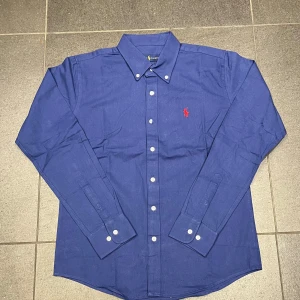 Blå skjorta från Ralph Lauren M - Klassisk blå skjorta från Ralph Lauren i bomull med button-down krage och vita knappar. Broderad röd logga på bröstet. Skjortan har lång ärm och rak passform, perfekt för en clean och stilren look.