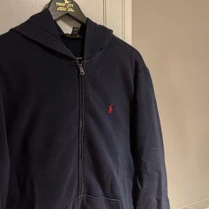 Mörkblå hoodie från Polo Ralph Lauren - Säljer en mörkblå hoodie från Polo Ralph Lauren med dragkedja och huva. Tröjan har det klassiska röda Polo-logot broderat på bröstet och två fickor framtill. Nypris ca 1200kr. Storlek XL, men passar även L. Hör av dig vid fler frågor. 