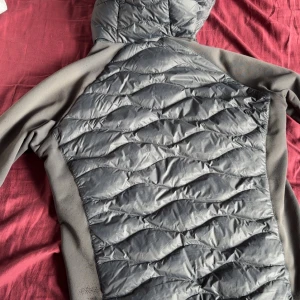 Grå quiltad Peak Performance jacka  - Snygg grå jacka från Peak Performance med quiltad framsida och huva. Jackan har dragkedja, bröstficka med zip och släta ärmar i stretchigt material. Perfekt för lager-på-lager och outdoor. Logga på bröstet och sportig look. 