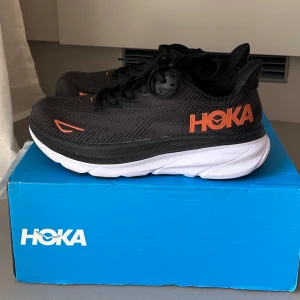 Hoka Clifton 9 svarta sneakers - Säljer ett par Hoka Clifton 9 sneakers i svart med orange detaljer och vit sula. Perfekta för dig som gillar att springa eller vill ha riktigt sköna skor till vardags. Aldrig använda då jag råkade beställa fel storlek