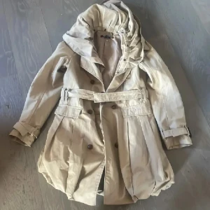 Beige trenchcoat med huva - Snygg beige trenchcoat  med stor huva, dubbelknäppning och bälte i midjan. Jackan har långa ärmar med spänndetaljer vid handlederna och en ballongformad nederkant som ger en cool siluett. Perfekt för vår och höst! Superfin