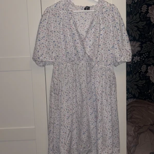 Blommig omlottklänning från PIECES - Supersöt vit omlottklänning från PIECES med små blommor i blått, rött och grönt. Klänningen har korta puffärmar, resår i midjan och v-ringning. 