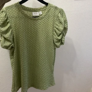 Grön topp med puffärmar från Vila - Säljer en ljusgrön topp från Vila med korta puffärmar och strukturerat tyg. Toppen har rund halsringning och en unik textur som ger en cool look. Perfekt för dig som vill sticka ut med en trendig och bekväm stil.