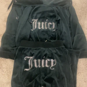 Juicy Couture - Mörkgrön kofta från Juicy Couture i mjuk velour med glittrande Juicy-text i strass på ryggen. Klassisk modell med huva och snörning, perfekt för en chill och trendig look. Matcha med byxorna för den ikoniska Juicy-stilen. Koftan är i storlek xxs, byxorna är i storlek xs.
