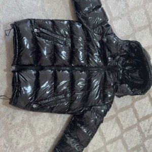 Moncler - Säljer en svart, glansig dunjacka från Moncler med huva och dragkedja framtill. Jackan har flera fickor med dragkedja och Moncler-logga på ärmen. Puffig modell med justerbara snören och snygga detaljer. Perfekt för kalla dagar. Har även NFC TAGG