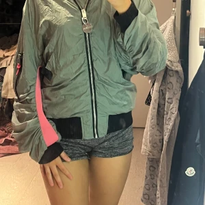 Grå bomberjacka från Diesel - Säljer en grå bomberjacka från Diesel med svarta muddar och dragkedja framtill. Jackan har en cool detalj med rosa insida på ena ärmen och en stor färgglad patch på insidan med text. Perfekt statement-plagg för dig som vill sticka ut.