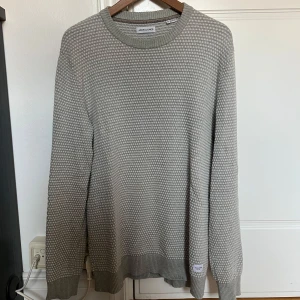 Grå stickad tröja Jack & Jones XL - Säljer en grå stickad tröja från Jack & Jones i storlek XL. Tröjan har ett diskret prickigt mönster, rund halsringning och ribbade muddar vid ärmslut och nederkant. Perfekt för lager-på-lager under höst och vinter. Tröjan är liten i storleken och sitter mer som L.
