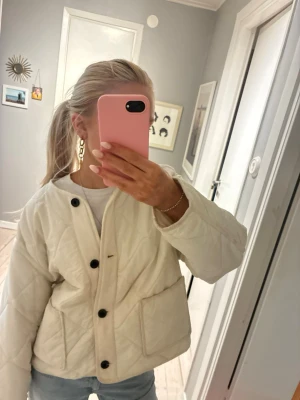 Vit Zara jacka - Säljer en quiltad beige jacka med svarta knappar och stora fickor framtill. Jackan har en rak och lite boxig passform, är långärmad och har ett stilrent, minimalistiskt uttryck. Perfekt för dig som gillar en clean och trendig look.