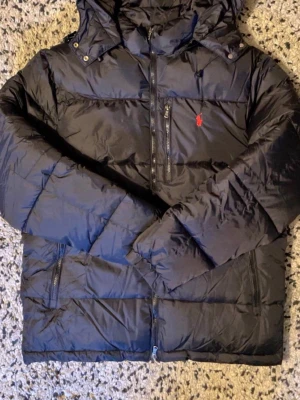 Svart pufferjacka från Polo Ralph Lauren - Säljer en svart pufferjacka från Polo Ralph Lauren med röd broderad logga på bröstet. Jackan har huva, flera dragkedjeförsedda fickor och quiltad design. Perfekt för kalla dagar och riktigt snygg till streetwear-stilen.