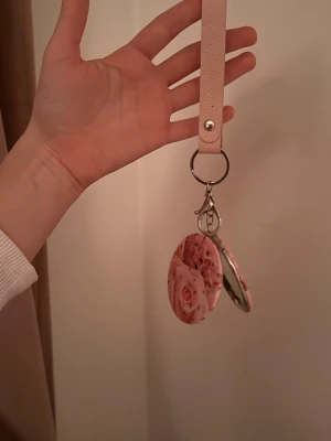 Rosa sminkspegel med blommor och nyckelring - Söt rund sminkspegel med rosa blommotiv och små glittriga stenar på locket. Spegeln har en praktisk nyckelring och en ljusrosa rem i konstläder, perfekt att hänga på väskan eller nyckelknippan. Smidig att ta med för snabba touch-ups.