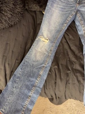 Blå bootcut jeans med slitningar - Säljer ett par blå bootcut jeans från Perfect Jeans med slitningar på låret och vid fickan fram. Jeansen har snygga kontrastsömmar och bakfickor med dekorativa sömmar och knappstängning. 