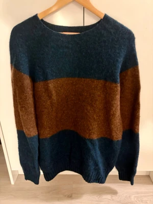 Randig stickad tröja i blått och brunt - Mysig stickad tröja med breda ränder i mörkblått och brunt. Tröjan har rund halsringning och långa ärmar. Perfekt för kyliga dagar och enkel att matcha med jeans. 100% ull 