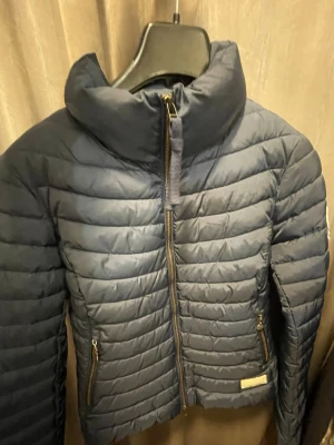 Mörkblå pufferjacka från Odd Molly - Snygg mörkblå pufferjacka/dunjacka från ikoniska Odd Molly med broderad logga på ärmen och guldfärgade dragkedjor. Jackan har hög krage, två fickor med dragkedja och coola detaljer; exempelvis ett dekorativt dragkedjehänge och coola detaljer i jackan. Perfekt för kyliga dagar. Säljs i befintligt skick men är i väldigt bra skick. Hör av dig vid intresse😊