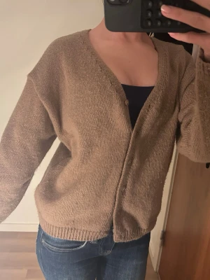 Beige stickad kofta med knappar - Mysig brun stickad kofta med v-ringning och knappar framtill. Koftan har en loose passform och långa ärmar, perfekt att slänga över en topp. Ribbstickade muddar vid ärmslut och nederkant ger en avslappnad vibe.