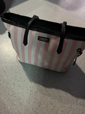 Randig shoppingväska Victoria's Secret - Säljer en stor shopping  väska från Victoria's Secret med klassiska rosa och vita ränder. Väskan har svarta handtag och detaljer i konstläder samt guldfärgade metalldetaljer på sidorna. Perfekt för dig som vill ha plats för allt och ändå se stylish ut.✨💕