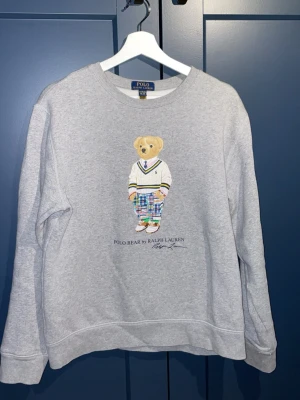 Grå tröja Polo Bear Ralph Lauren - Grå tröja från Polo Ralph Lauren med Polo Bear-tryck framtill. En gosig tröja med skönt material inuti som är i fint skick! Är i storlek XL/18-20 vilket motsvarar S i vuxen storlek, säljs för att den blivit för liten, hör av er vid funderingar!