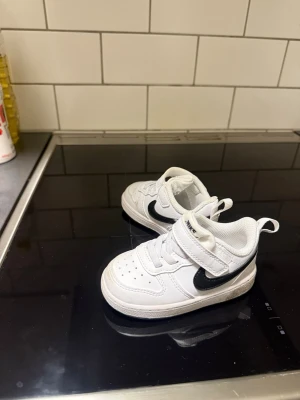 Vita Nike sneakers för barn - Säljer ett par vita Nike sneakers för barn med svart swoosh på sidan. Skorna har kardborreband och elastiska skosnören för enkel påtagning. Ovandelen är i syntetmaterial och sulan är svart undertill. Klassisk och sportig design som passar till det mesta.