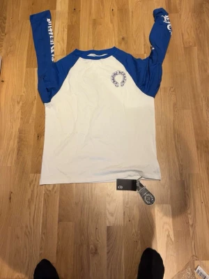 Chrome Hearts blå/vit långärmad long sleeve  - Säljer en långärmad t-shirt från Chrome Hearts med vita och blå kontraster. Stor CH-logga på ryggen och tryck längs båda ärmarna. Klassisk baseballmodell med rund hals och coola detaljer. Perfekt för dig som gillar streetwear och vill sticka ut.