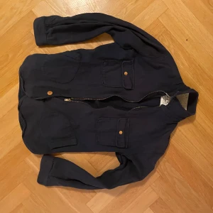 Mörkblå overshirt från Gooseberry Lay & Co - Säljer en mörkblå overshirt från Gooseberry Lay & Co i 100% ekologisk bomull. Jackan har två bröstfickor med knapp, dragkedja framtill med en knapp längs ner och klassisk krage. Perfekt lager-på-lager-plagg med enkel och stilren look. Knappen är lite sliten men annars inga riktiga problem. Skriv för detaljer.