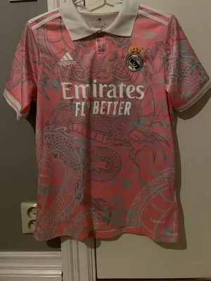 Rosa Real Madrid adidas tröja S - Unik rosa Real Madrid-tröja från adidas i storlek S. Tröjan har ett stort drakmönster i grått och vitt, vit krage och vita detaljer på axlarna. Tillverkad i lätt och ventilerande syntetmaterial, perfekt för fotboll eller streetwear.