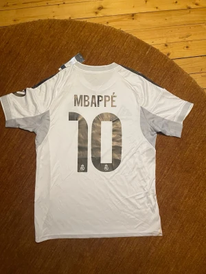 Real Madrid Mbappé tröja Adidas M - Säljer en vit Real Madrid matchtröja från Adidas med Mbappé och nummer 10 på ryggen. Tröjan har svarta detaljer, broderat klubbmärke, HP-logga på ärmen och gula linjer längs sidorna. Slim fit och tillverkad i polyester, perfekt för fotbollsfans.