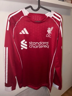 Långärmad Liverpool tröja med Isak - (Använt 1 gång) Säljer en röd Liverpool matchtröja från Adidas med vita detaljer, lång ärm och tryck fram och bak. På bröstet finns LFC-logga och sponsor, på ryggen står det ISAK och nummer 9. Tröjan är i slim fit och tillverkad i polyester.