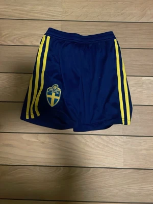Svenska fotbollslandslaget shorts - Mörkblå shorts från Adidas med Sveriges landslagsemblem på ena benet och klassiska gula ränder längs sidorna. Perfekta för fotbollsträning eller match. Tillverkade i lätt och ventilerande polyester.