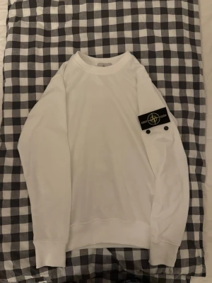 Vit Stone Island sweatshirt - Vit sweatshirt Stone Island med klassisk patch på vänster ärm. Tröjan har rund halsringning, ribbade muddar och är tillverkad i mjuk bomull. Perfekt för dig som gillar clean och stilren streetwear.