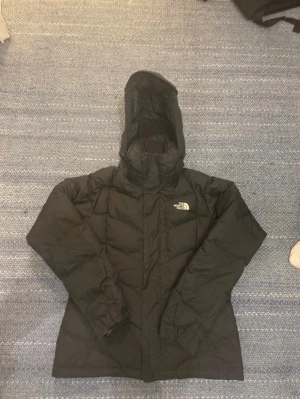 Svart dunjacka från The North Face - Säljer en svart dunjacka från The North Face med huva och dragkedja. Jackan har diskret mönstrat innerfoder och logga broderad på bröstet. Perfekt för kalla dagar och riktigt skön att bära. Passar dig som vill ha en varm och stilren jacka.