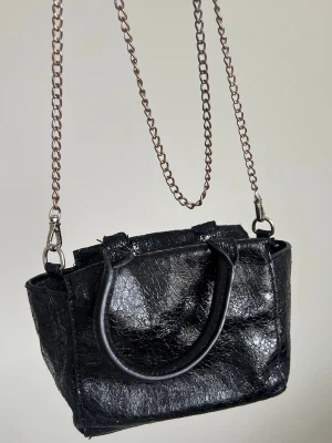 Väska  - Liten svart väska med glansig, krackelerad yta. Väskan har både handtag och en lång metallkedja som axelrem, samt dragkedja upptill. Mettalkedjan är något missfärgad på ställen. Från Nelly 🖤