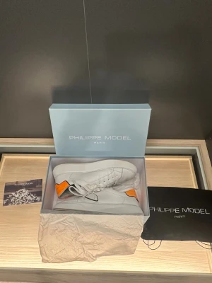 Philippe Model skor - Säljer ett par stilrena vita Philippe Model sneakers i skinn med en snygg orange detalj på hälen. Skorna har rund tå, vita snören och en platt sula. Klassisk logga på sidan och kontrastkant i svart. Perfekta för dig som gillar premium och clean look.