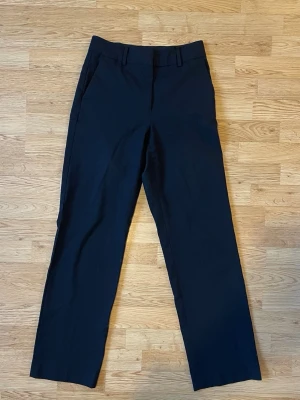 Svarta kostymbyxor Stockholm LM - Snygga svarta kostymbyxor med raka ben och klassiska fickor fram. Byxorna har hällor för bälte och en enkel bakficka med knappdetalj. Perfekta för en clean och stilren look.