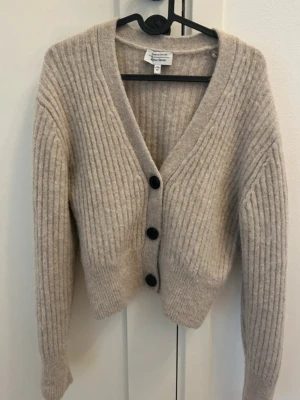 Beige ribbad kofta från & Other Stories - Supermysig beige kofta från & Other Stories med ribbat stickat material och stora svarta knappar framtill. 
