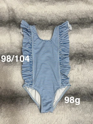 Blåvit randig baddräkt med volanger - Säljer en söt blåvit randig baddräkt med volangdetaljer längs axelbanden och sidorna. Perfekt för strand eller poolhäng. 🌸 Fint använt skick. Storlek 98/104