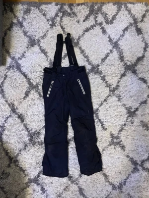 Svarta täckbyxor från Everest 146 - Svarta täckbyxor från Everest med justerbara hängslen och vita dragkedjor framtill. Byxorna är tillverkade i slitstarkt syntetmaterial och har en rak passform som passar perfekt för vinterns alla äventyr. Märket syns diskret på benet. Storlek 146