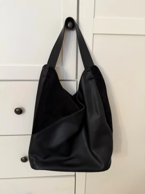 Svart axelväska från DonDonna - Köpt för 699kr och helt oanvänd! 🥰Snygg svart axelväska från DonDonna i en mix av slätt skinn och mocka. Väskan har bred axelrem och en cool asymmetrisk design som ger en modern vibe. Perfekt storlek för att få plats med allt du behöver och funkar till alla outfits!! 