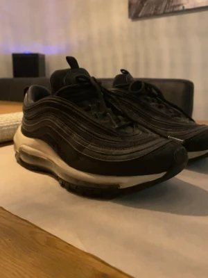 Svarta Nike Air Max 97 sneakers - Säljer ett par svarta Nike Air Max 97 sneakers med ikoniska vågiga linjer och synlig Air-enhet i sulan. Skorna har snörning, rund tå och en mix av mesh och syntetmaterial för en sportig look. Perfekta för dig som gillar streetwear och vill ha bekväma skor till vardags.