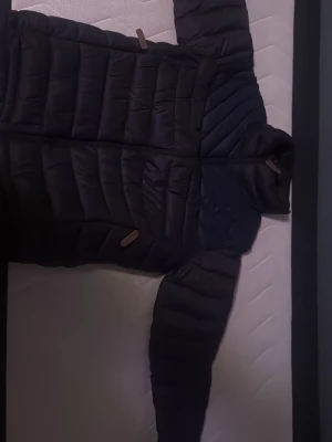 Svart pufferjacka med dragkedja - Säljer en svart pufferjacka med quiltad design och långa ärmar. Jackan har dragkedja framtill och detaljer i form av bruna läderflikar på dragkedjorna. Perfekt för kyliga dagar och riktigt skön att bära.