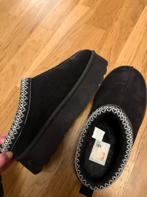 Svarta mockatofflor med mönstrad kant - Mysiga svarta tofflor i mocka med tjock sula och dekorativt broderad kant i vitt. Insidan är fodrad för extra värme och komfort. Perfekta för kalla dagar. 