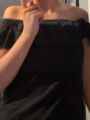 Svart offshoulder topp med volang - Säljer en svart offshoulder topp med bred volangkant runt axlarna. Toppen är ärmlös och har en snygg broderad detalj på volangen. Perfekt till jeans eller kjol för en trendig look.