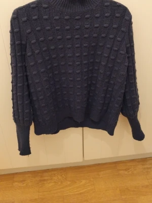 Mörkblå stickad polotröja - Säljer en mörkblå stickad polotröja med upphöjt prickigt mönster över hela tröjan. Tröjan har lång ärm och hög krage som ger en mysig känsla. Perfekt för kyliga dagar och enkel att styla med jeans eller kjol.