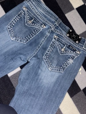 Blå bootcut jeans med stjärnlapp - Säljer ett par blå bootcut jeans från Miss Me med kontrastsömmar, snygga detaljer på bakfickorna och en stor stjärnpatch på ena benet. Jeansen har dekorativa knappar och cool tvättad look. Perfekta för dig som vill sticka ut lite extra!