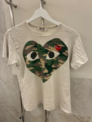 Comme des Garçons Play camo t-shirt - Säljer en vit Comme des Garçons Play t-shirt i bomull med stort hjärta i camouflageprint och ögon på bröstet. Klassisk rund hals och korta ärmar. Liten röd hjärtdetalj med ögon broderad på hjärtat. Perfekt statement-plagg för streetwear.
