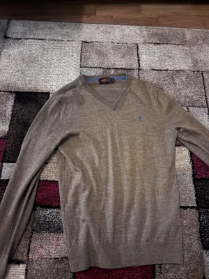 Beige merinoullströja från Morris - Säljer en beige V-ringad tröja från Morris i 100% merinoull. Tröjan har lång ärm, ribbade muddar och en liten blå broderad logga på bröstet. Perfekt för dig som gillar stilrena och klassiska plagg.