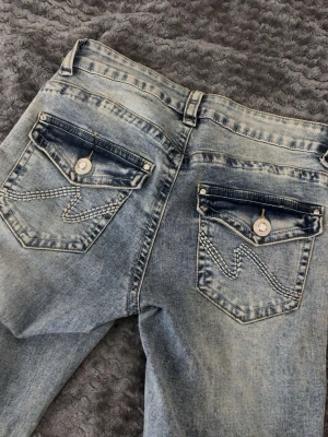  ljusblå Bootcut jeans med detaljer - Säljer ett par ljusblå jeans från Missay med snygg tvätt och kontrastsömmar. Bakfickorna har lock och dekorativa sömmar samt knappar med bling. Klassisk femficksmodell och dragkedja framtill. Materialmix med bomull, polyester, rayon och elastan.