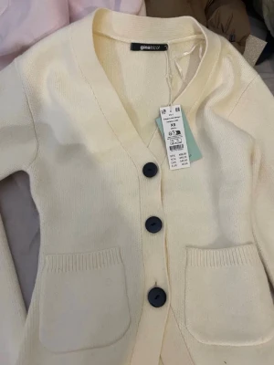 Beige kofta från Gina Tricot - Mysig beige kofta från Gina Tricot med stora svarta knappar och två fickor framtill. Koftan har en djup v-ringning och är stickad i mjukt material, perfekt för lager på lager under kyliga dagar.