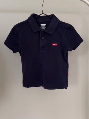 Svart pikétröja från Levi's - Klassisk svart pikétröja från Levi's med korta ärmar och krage. Tröjan har tre knappar framtill och en liten röd Levi's-logga på bröstet. Perfekt till jeans eller shorts för en avslappnad stil.